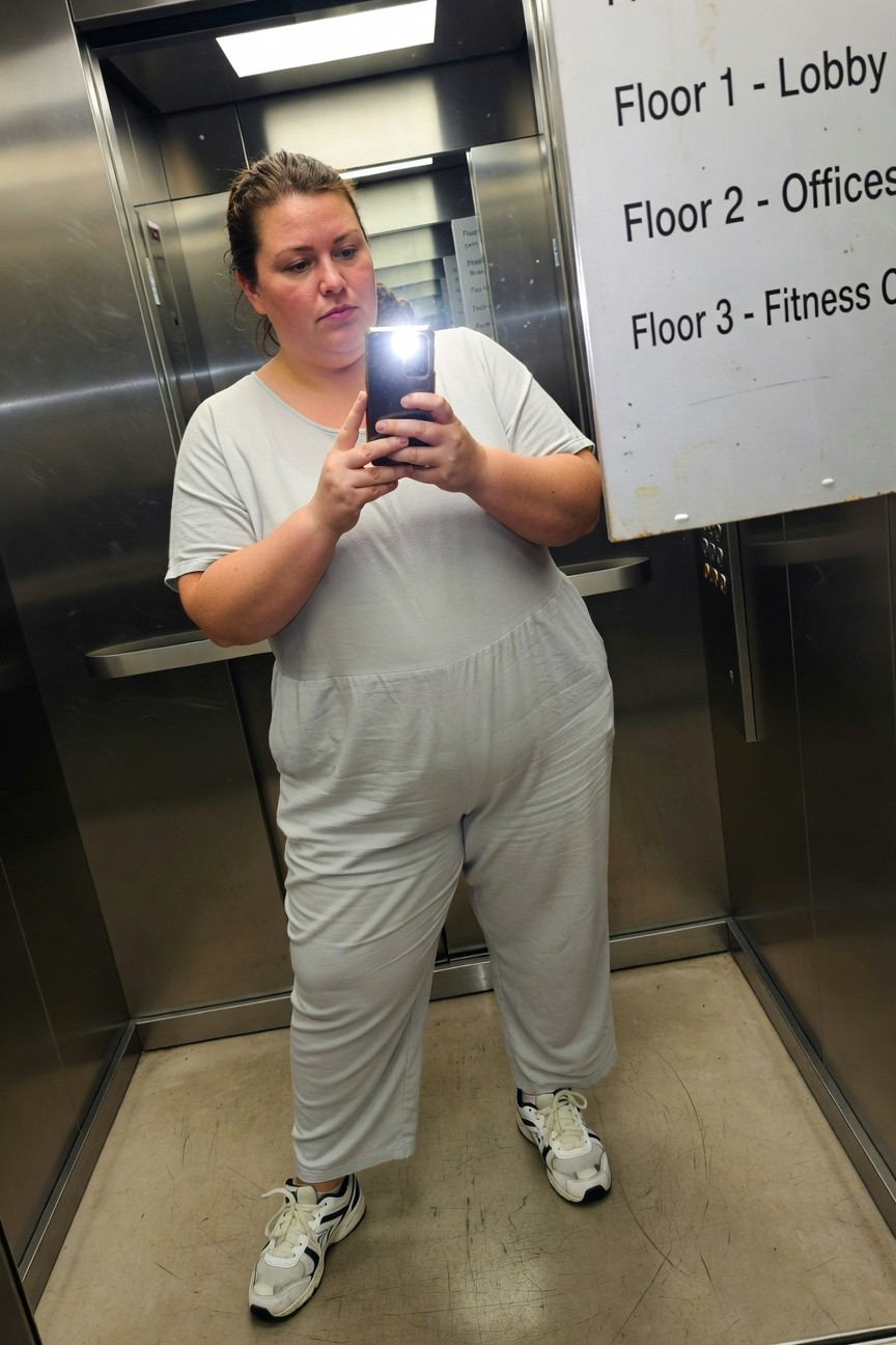 Plus-size woman in a loose light gray short-sleeve wide-leg jumpsuit and white chunky sneakers, taking a selfie in a steel elevator with floor directory sign visible