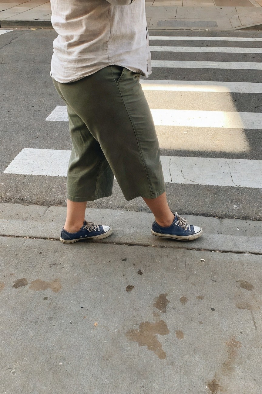 Plus-size woman from behind walking across a zebra crossing in a loose beige linen shirt, olive green wide-leg cropped pants ending mid-calf, and worn navy Converse low-top sneakers on a city street.