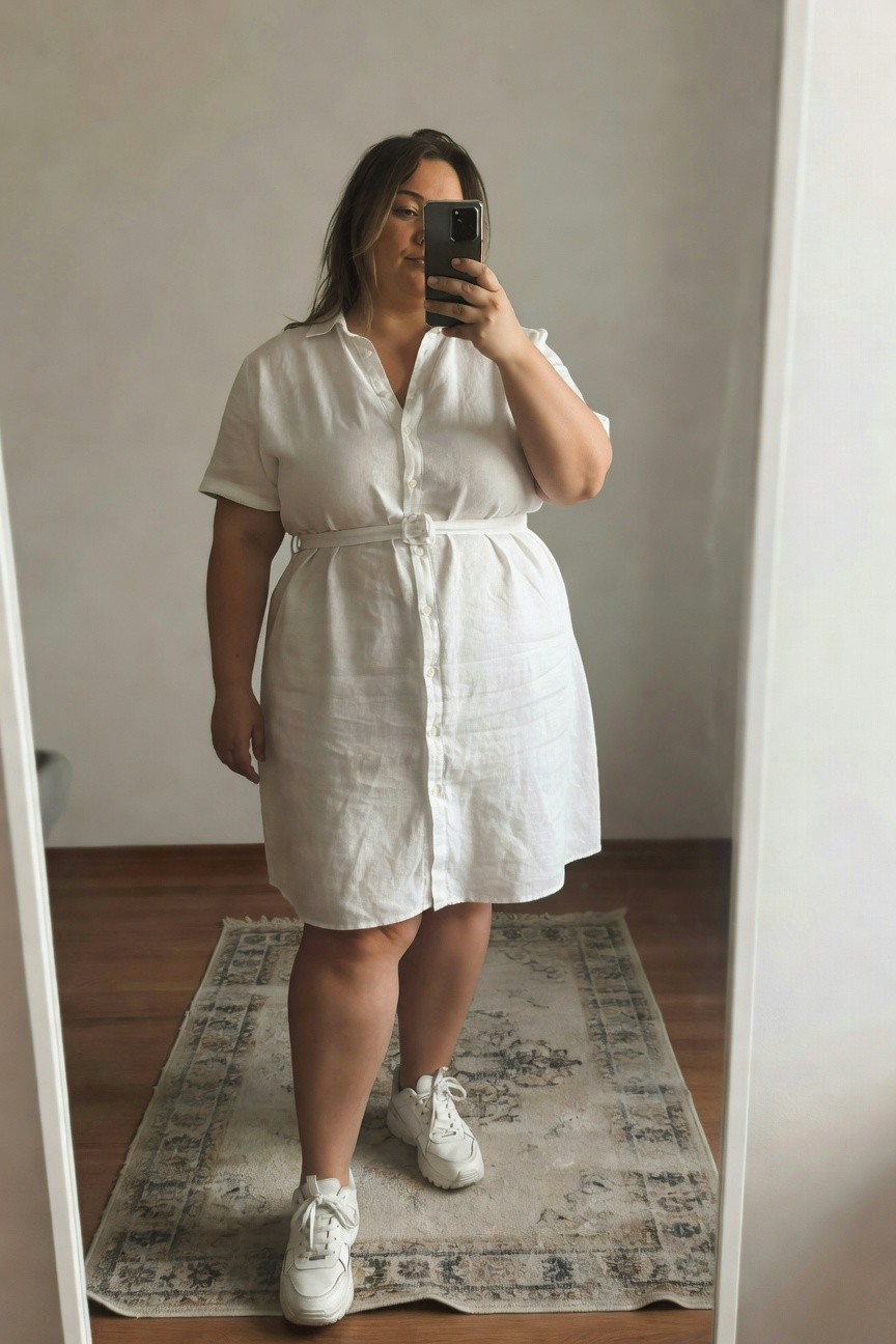 Plus-size woman taking a mirror selfie in a white short-sleeve linen button-front shirt dress with waist belt, paired with white chunky sneakers on a wooden floor with rug