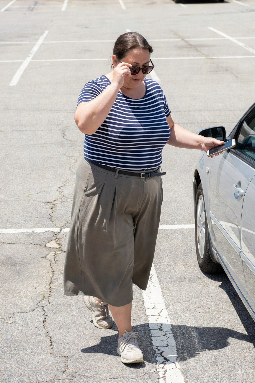 Plus-size woman in navy white striped short-sleeve tee, gray pleated wide-leg pants with black belt, white sneakers, sunglasses on head, holding phone near silver car in parking lot