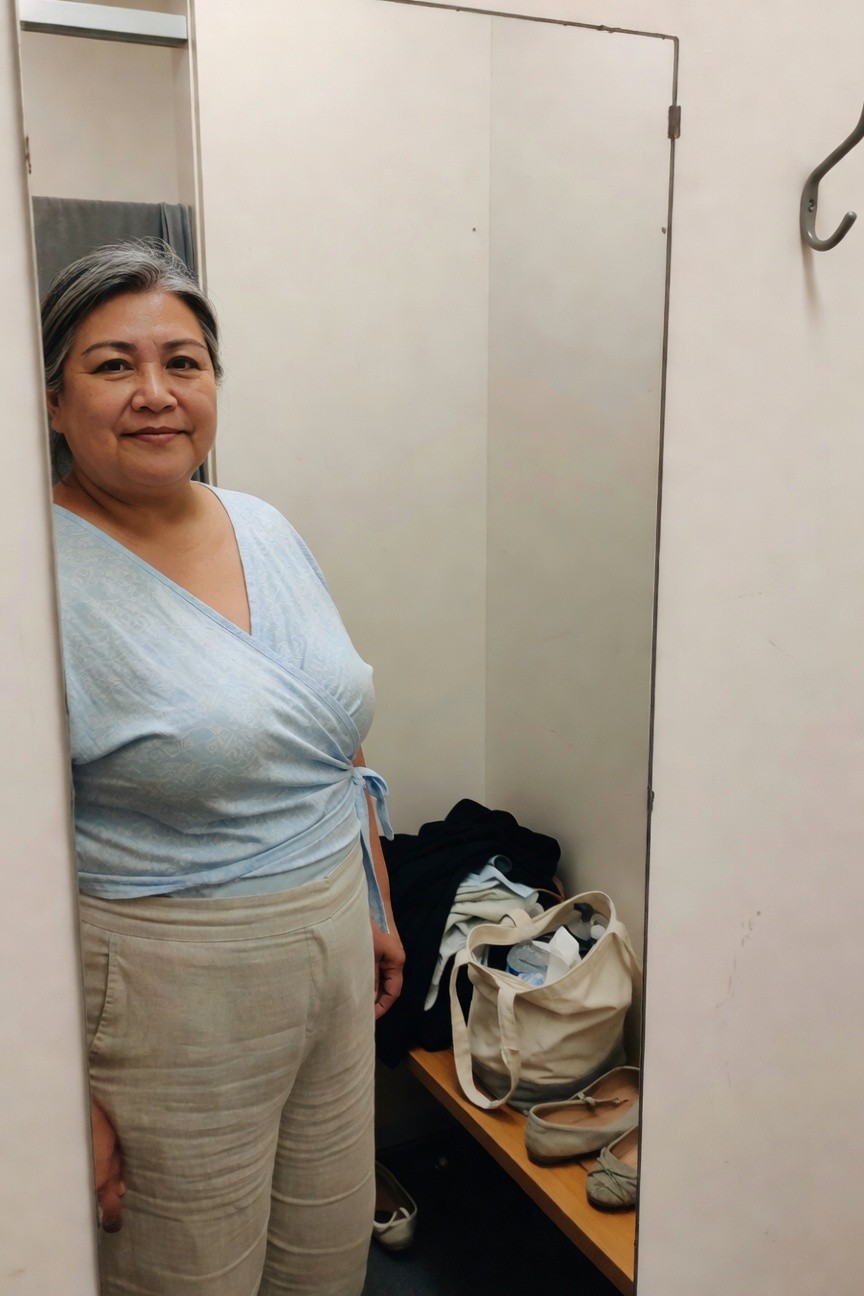 Plus-size woman over 40 smiling in changing room mirror selfie, wearing light blue patterned wrap blouse with deep v-neck and side tie over beige wide-leg linen pants, clothes bag and nude flat shoes on bench nearby