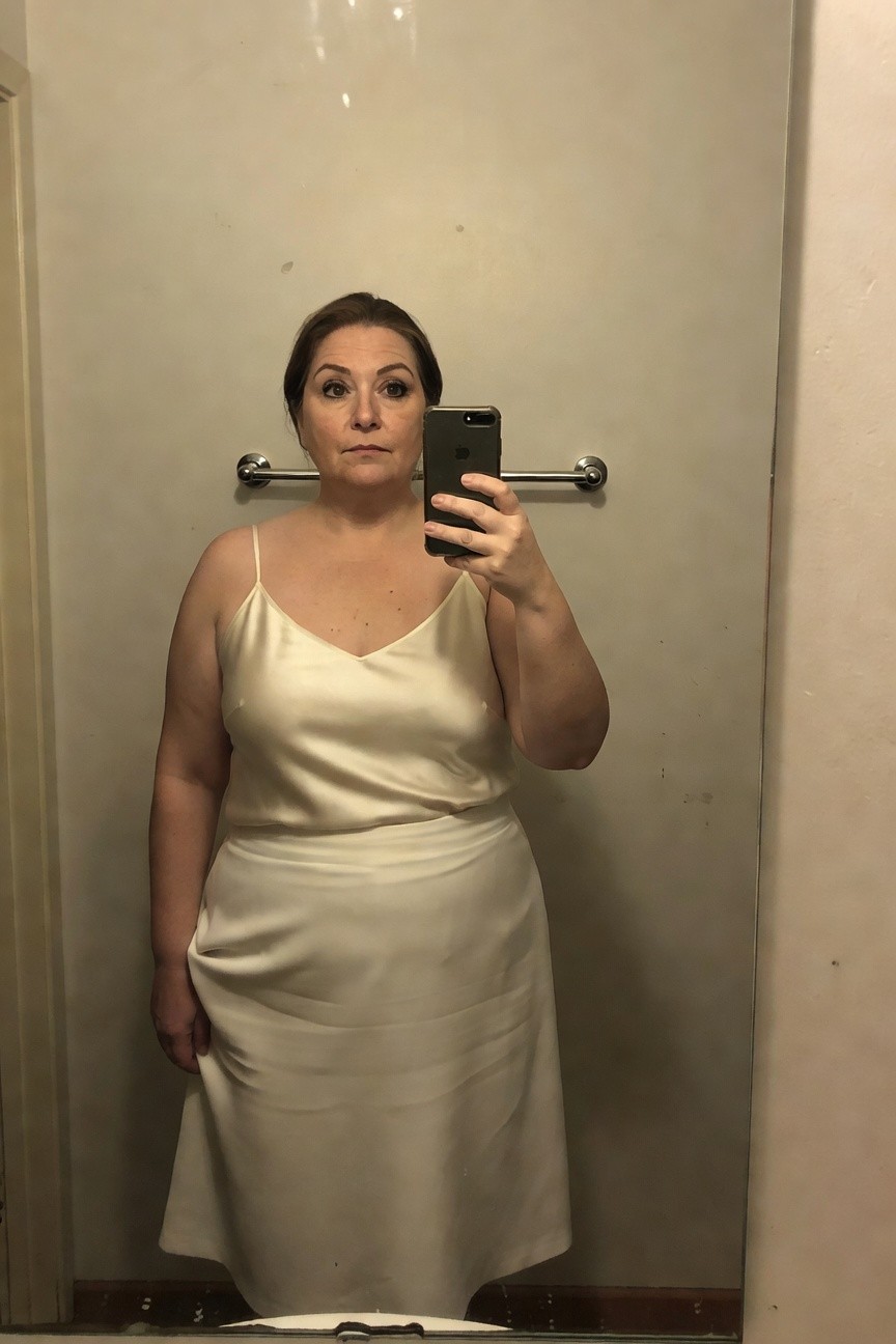 Plus-size woman over 40 in a cream satin spaghetti-strap slip dress with V-neck and A-line midi skirt, taking a mirror selfie in a bathroom.