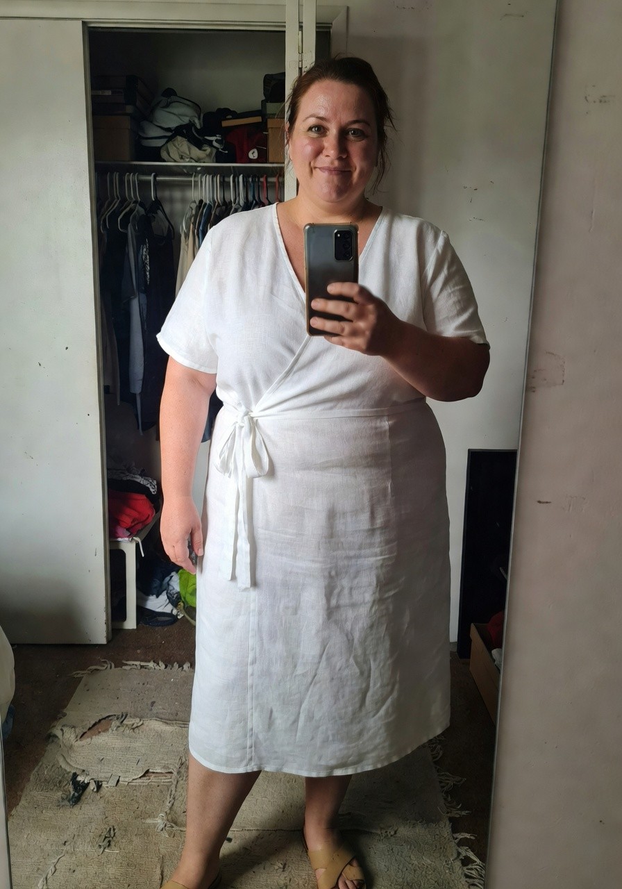 Plus-size woman over 40 in a white linen short-sleeve wrap midi dress with waist tie, posing in front of mirror wearing tan slide sandals, bedroom setting.