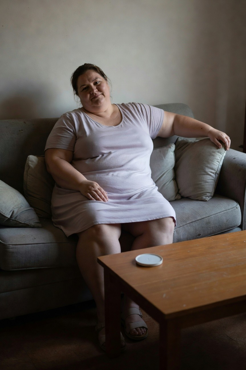 Plus-size woman over 40 seated on a gray couch in a loose pale pink sleeveless knee-length shift dress and beige strappy flat sandals, hands relaxed in lap