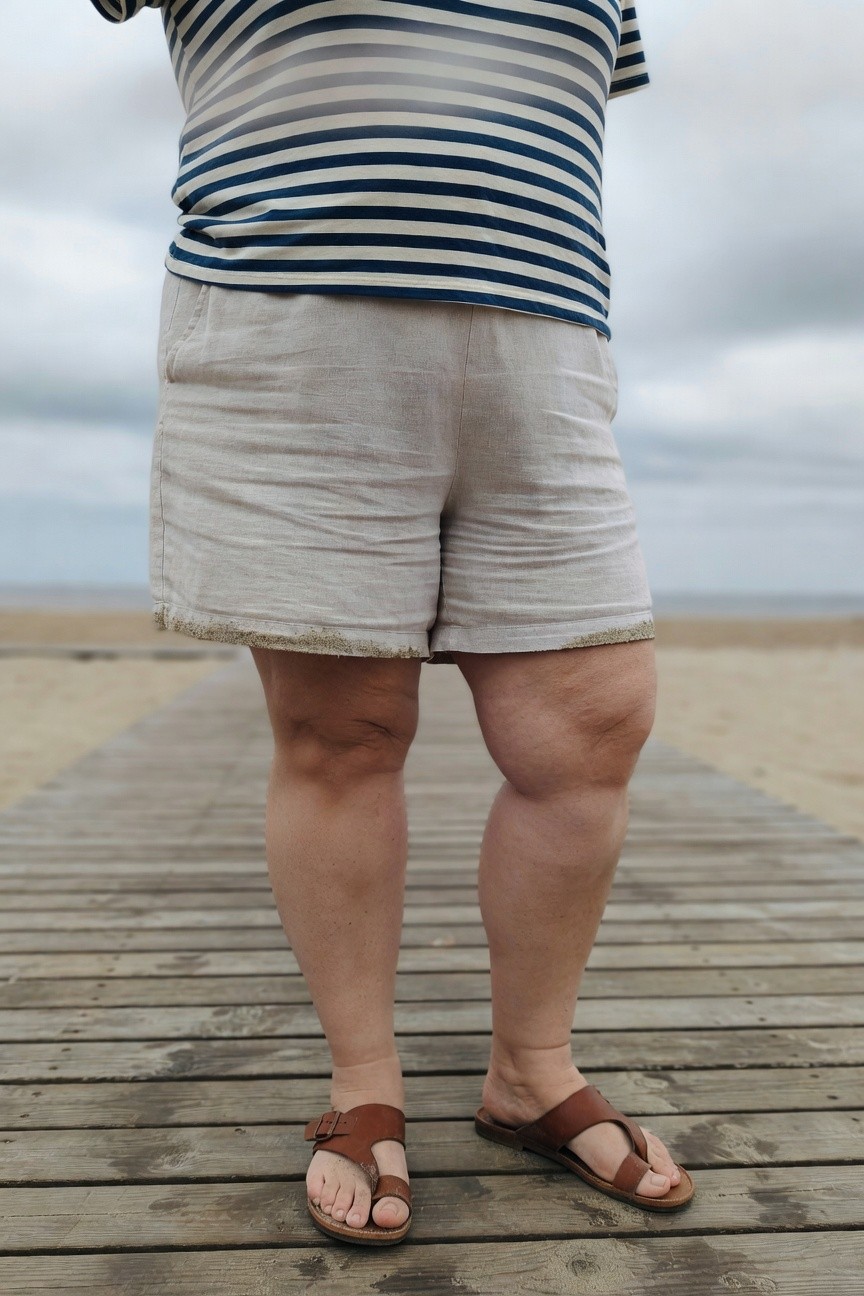 Plus-size woman over 40 in a navy and white horizontal striped short-sleeve tee tucked into high-waisted beige linen mid-thigh shorts with sand on the hems, wearing brown leather thong strap sandals, standing on a wooden pier by the beach