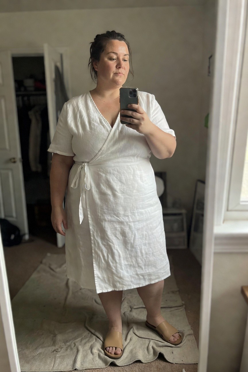 Plus-size woman over 40 in a white linen short-sleeve wrap midi dress with waist tie, posing in front of mirror wearing tan slide sandals, bedroom setting.