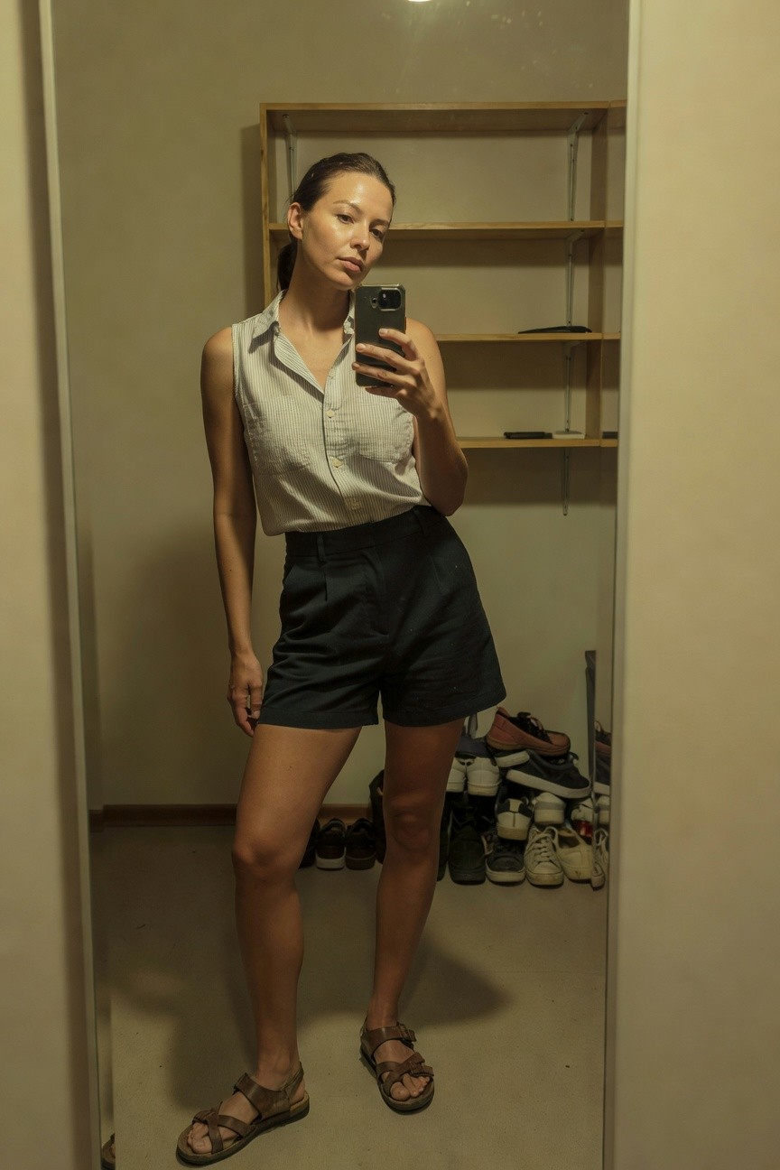 Mirror selfie of a woman in a light sleeveless button-up shirt, dark high-waisted pleated shorts, and tan strappy sandals, standing in a closet with shoe shelves in the background