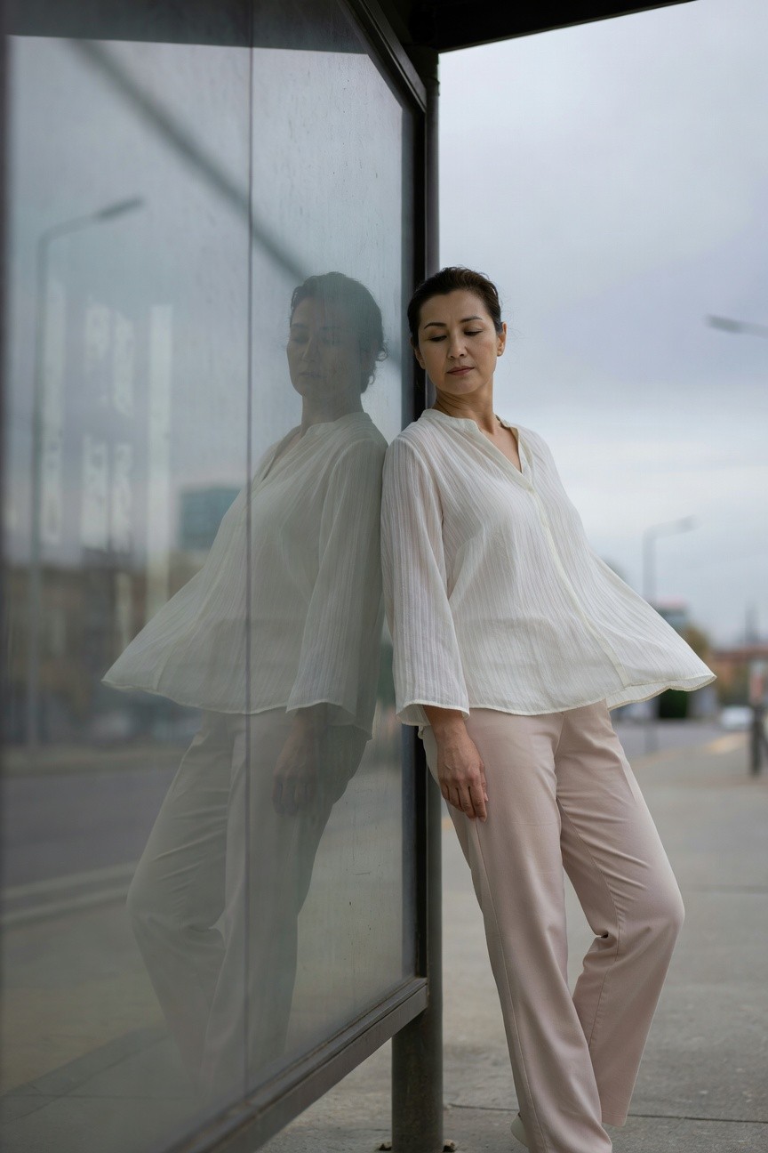 Asian woman in her 40s leaning against a glass bus shelter, wearing a textured white V-neck blouse with flared sleeves and wide-leg beige pants, her reflection visible in the glass, overcast urban street background
