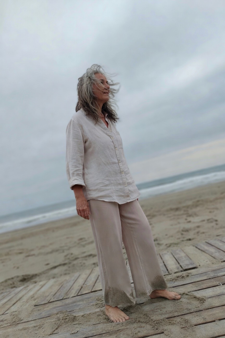 Older woman with gray wavy hair wearing a loose beige linen button-up shirt with rolled sleeves and matching wide-leg linen pants, standing barefoot on a wooden boardwalk with sand on the pant hems