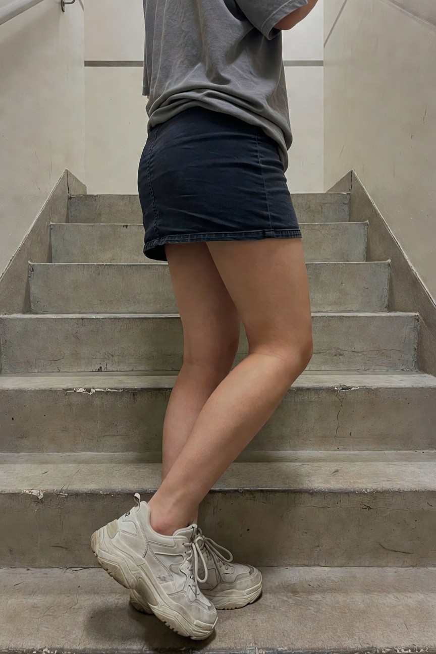 Woman viewed from behind on concrete stairs, wearing loose gray oversized t-shirt with one sleeve pushed up, short dark blue denim mini skirt, and chunky white platform sneakers with thick soles.