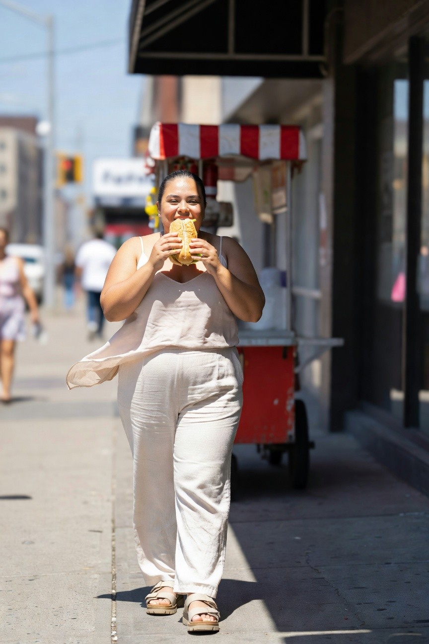 Plus size woman wearing flowy cream tank top, white wide leg linen pants, strappy sandals, and gold earrings, holding a corn cob while standing on a city sidewalk