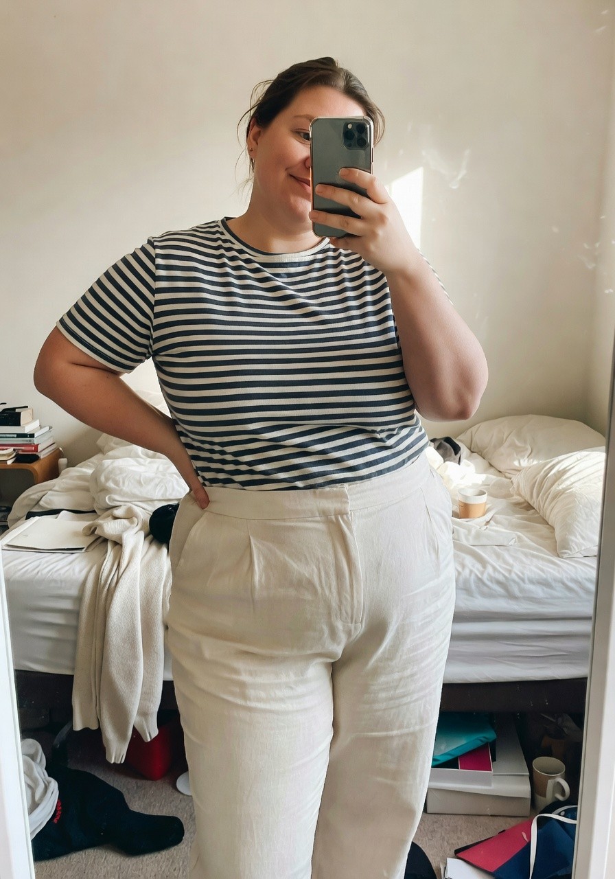 Plus-size woman in a white and navy horizontal striped short-sleeve t-shirt tucked slightly into high-waisted cream linen wide-leg pants, posing with hand on hip in front of a mirror selfie style