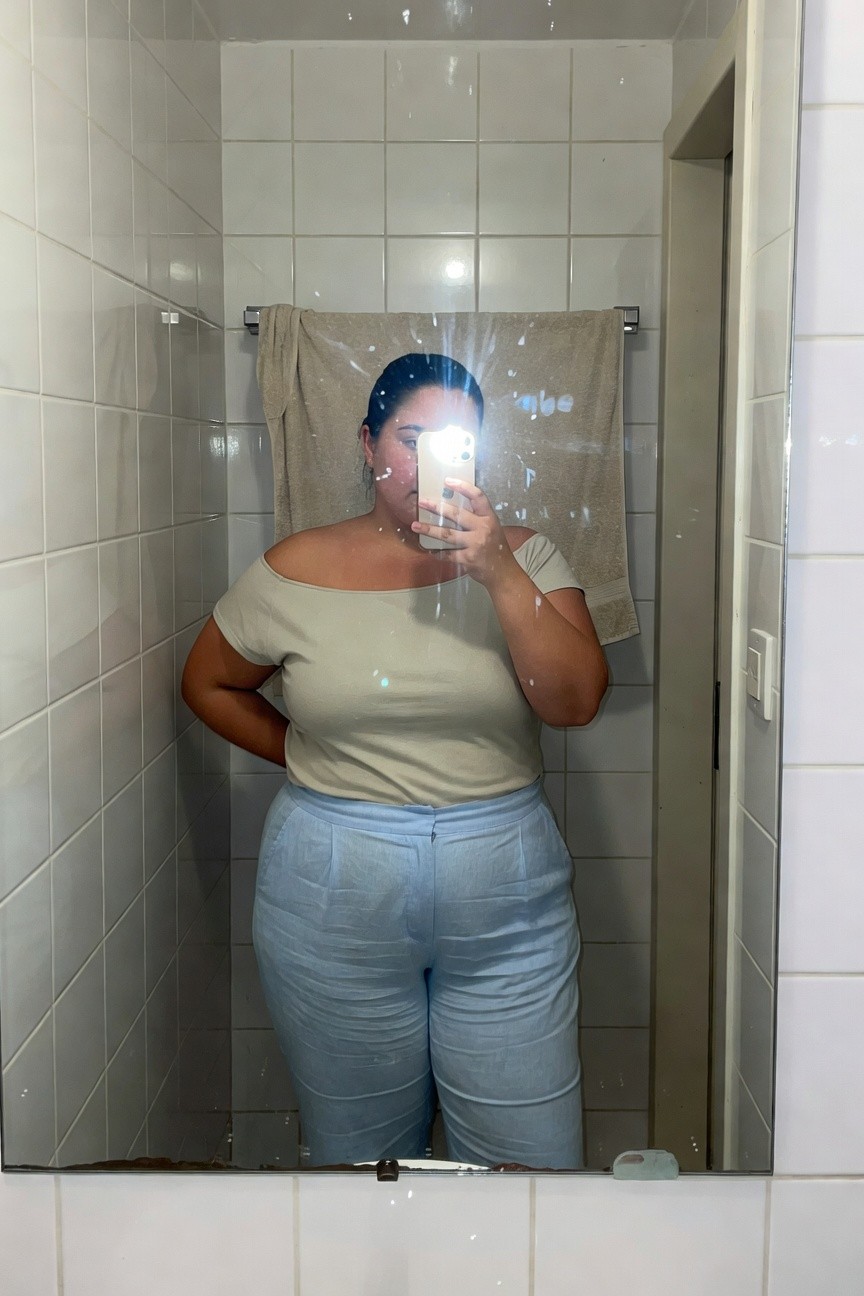 Plus-size woman taking a mirror selfie in a tiled bathroom, wearing a beige off-shoulder short-sleeve top and high-waisted light blue wide-leg linen pants, hands on hips with phone in one hand