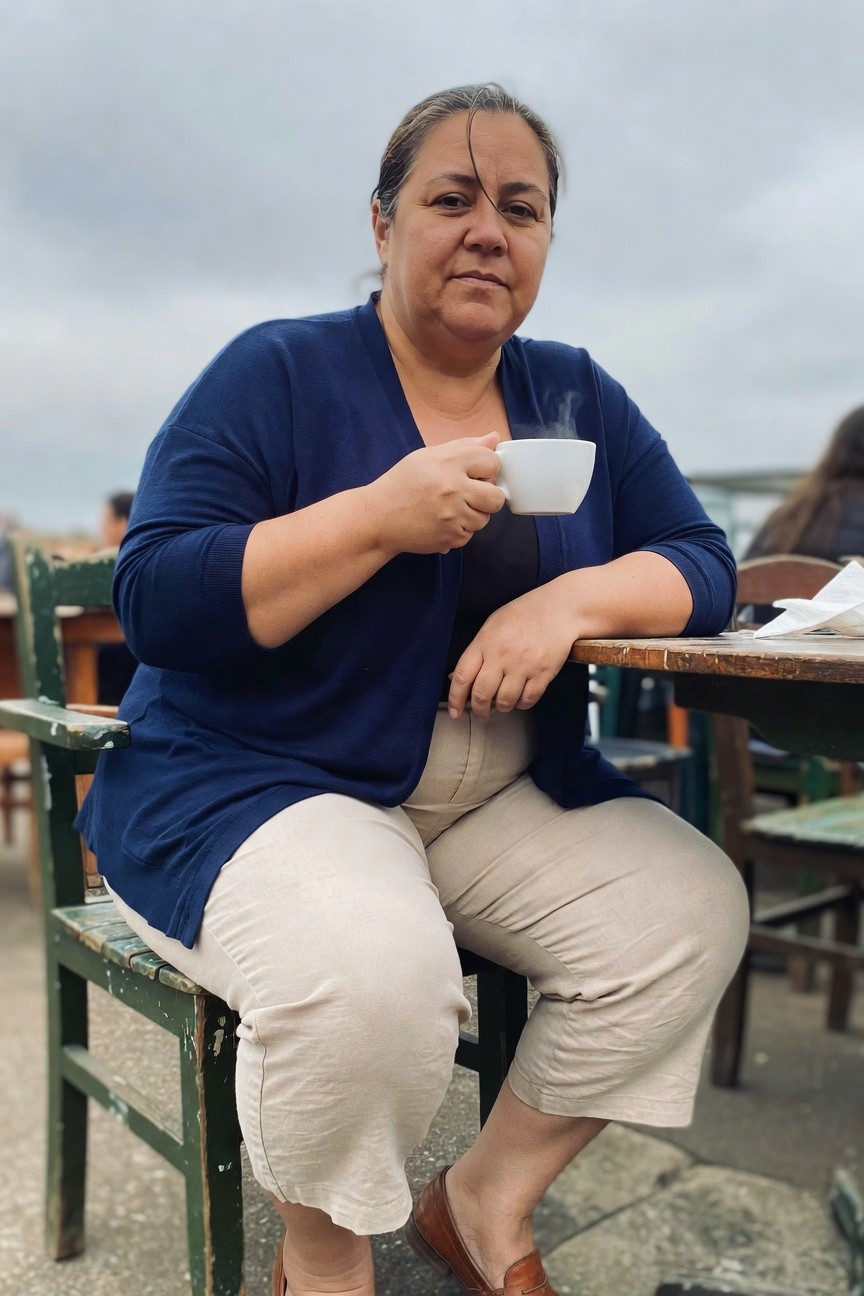 Plus-size woman seated outdoors at a wooden table, wearing an open navy blue long-sleeve cardigan over a dark top, paired with loose beige linen pants cropped at the ankle and brown loafers, holding a white mug of steaming drink