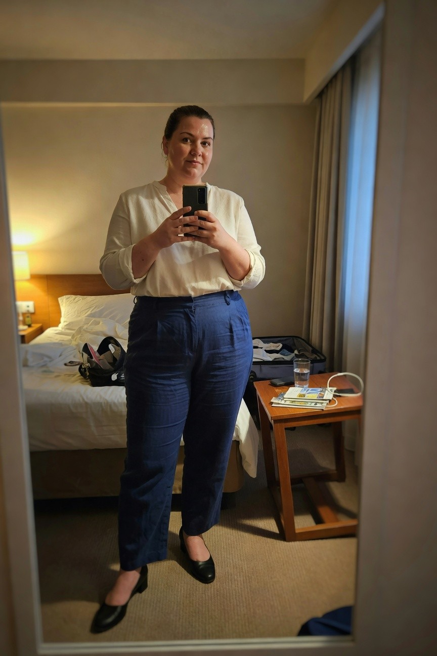 Plus-size woman in a mirror selfie wearing a white long-sleeve V-neck blouse tucked into high-waisted navy wide-leg linen pants and black low-heel flats, standing in a hotel room with bed and suitcase in background