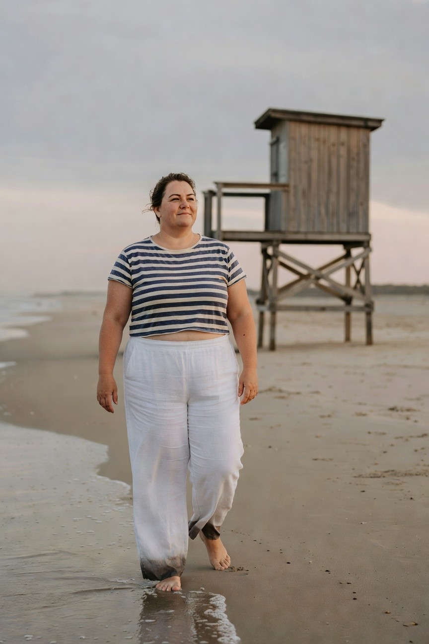 Plus-size woman with curly hair smiling while walking barefoot in shallow beach water, wearing a navy and white striped short-sleeve crop top and wide-leg white linen pants with soft hems, wooden lifeguard tower in background