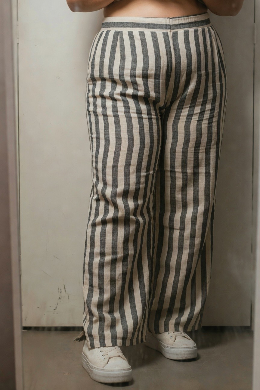 Plus-size woman in low-rise wide-leg black-and-cream striped linen pants showing midriff, hands on hips in front of mirror, wearing white sneakers on tiled floor