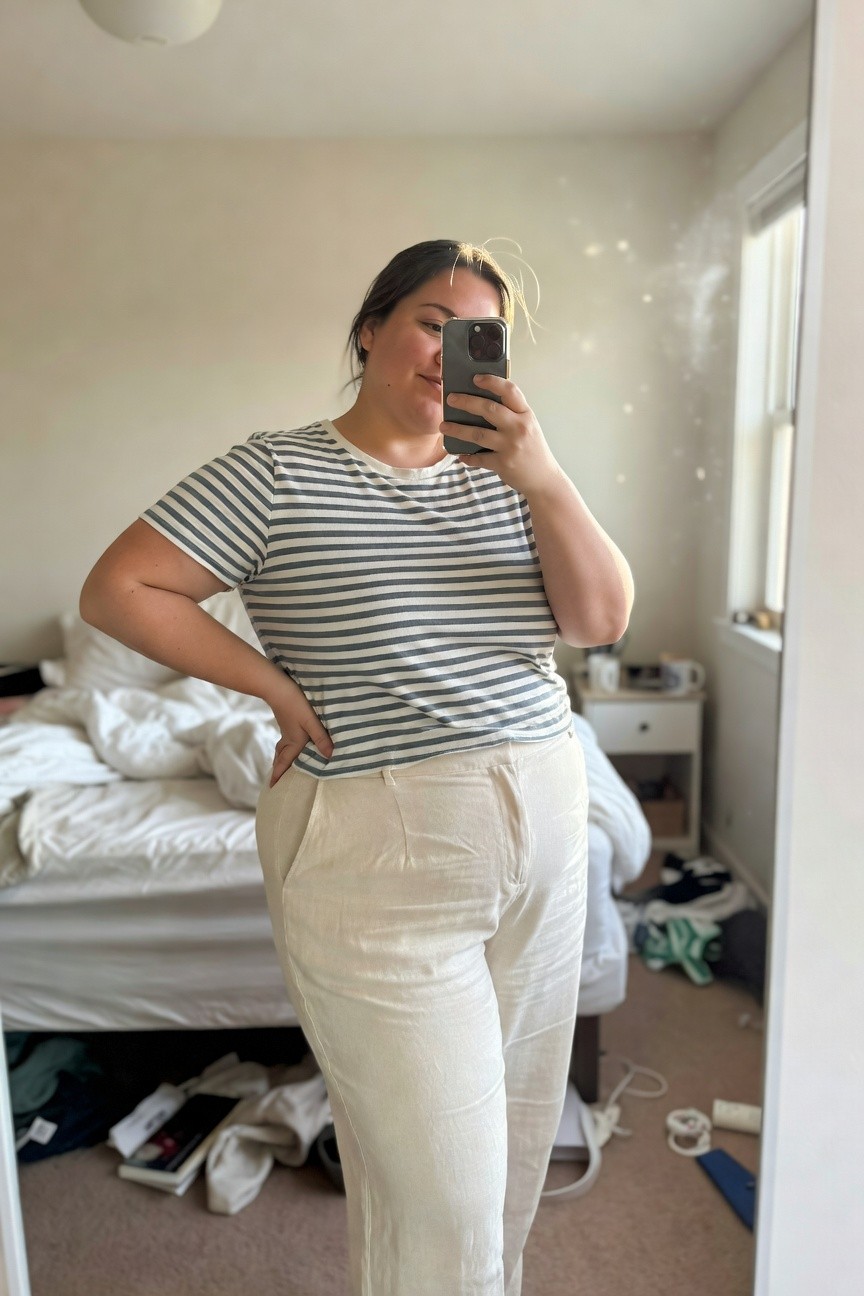 Plus-size woman in a white and navy horizontal striped short-sleeve t-shirt tucked slightly into high-waisted cream linen wide-leg pants, posing with hand on hip in front of a mirror selfie style
