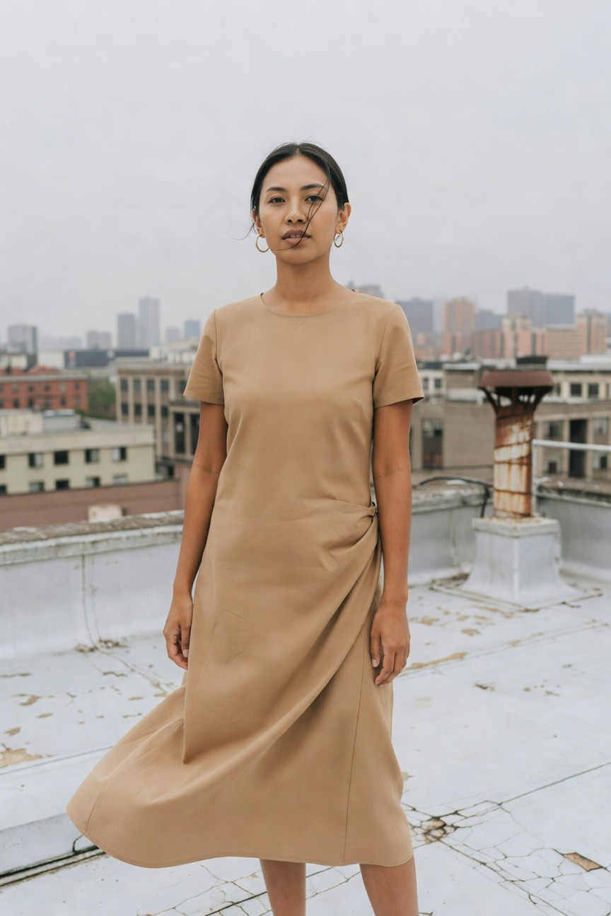 Woman in a light beige short-sleeve wrap midi dress with twisted waist detail, gold hoop earrings, standing on a rooftop against an urban skyline