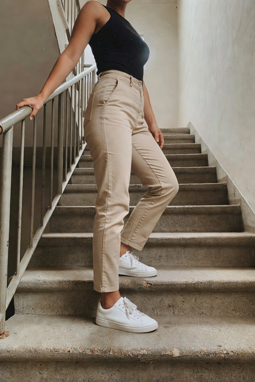 Woman leaning on metal stair railing, wearing black sleeveless ribbed tank top, high-waisted straight-leg khaki pants, and white leather sneakers, one foot on stair step