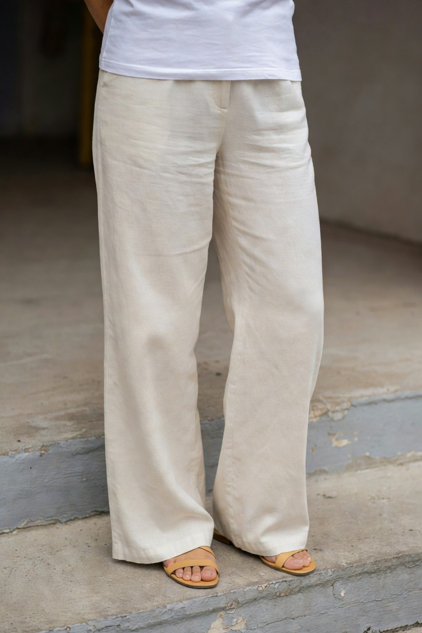 Woman in white short-sleeve t-shirt tucked slightly into high-waisted cream linen wide-leg pants with tan strappy flat sandals, posed sideways on concrete steps