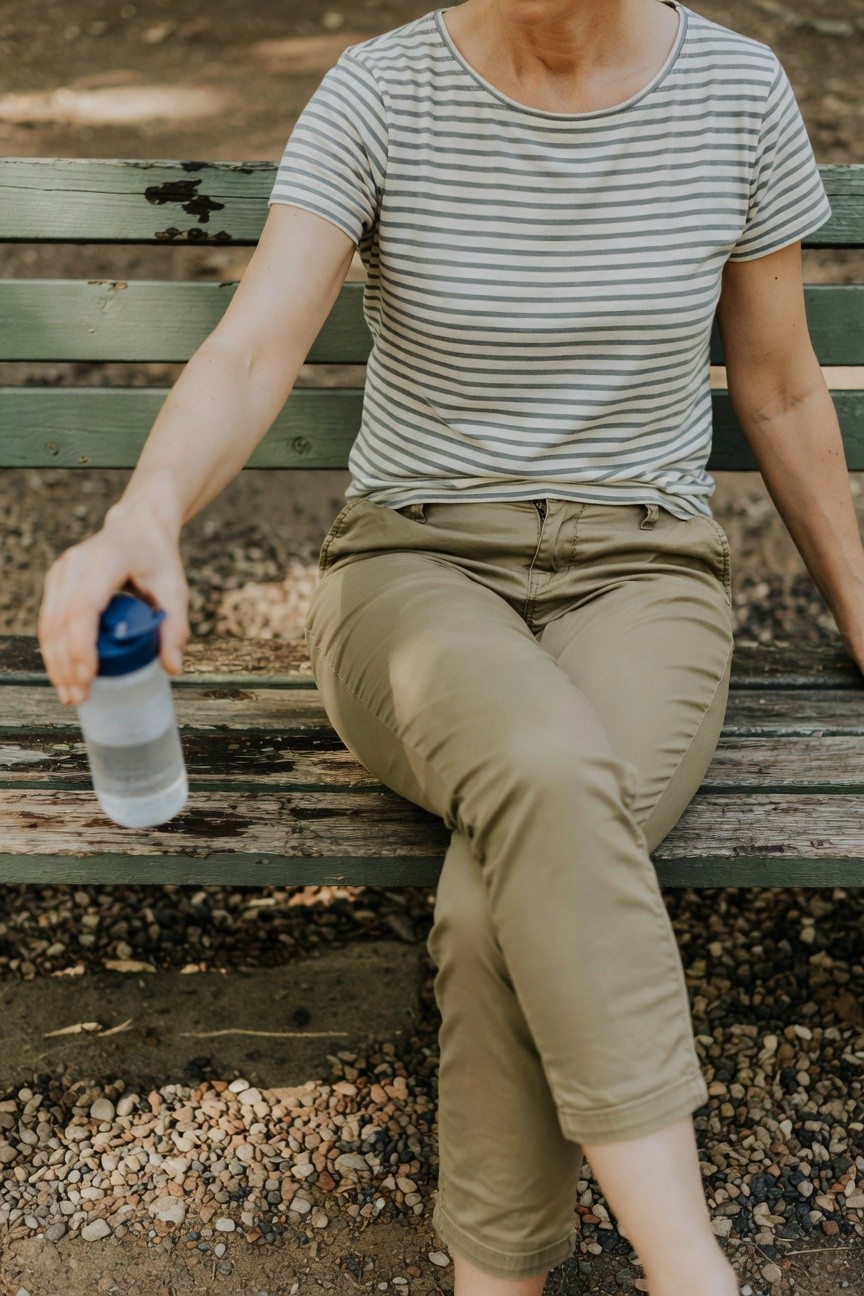 Mature woman sitting on wooden bench in a striped short-sleeve t-shirt and slim khaki pants, holding a blue-capped water bottle, legs crossed barefoot on gravel ground