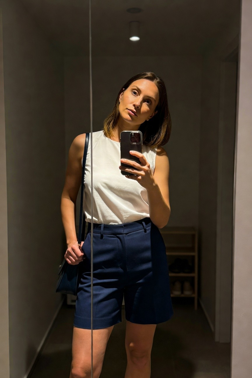 Woman taking mirror selfie in white sleeveless tank top, navy tailored shorts, black leather shoulder bag, in modern hallway with shoe shelves