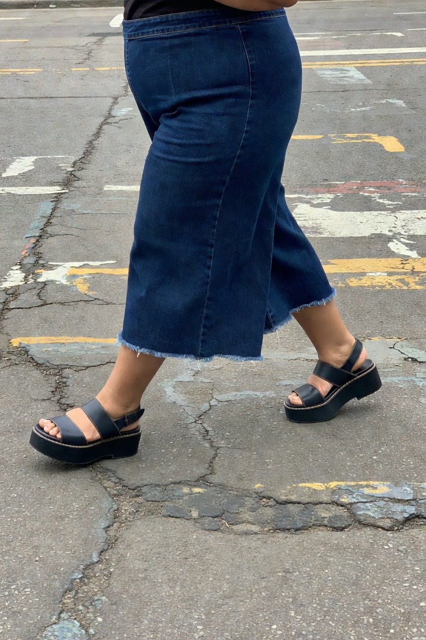 Side view of plus-size woman in black tank top, high-waisted dark blue wide-leg jeans with frayed ankle hem, and black double-strap platform sandals, walking on pavement