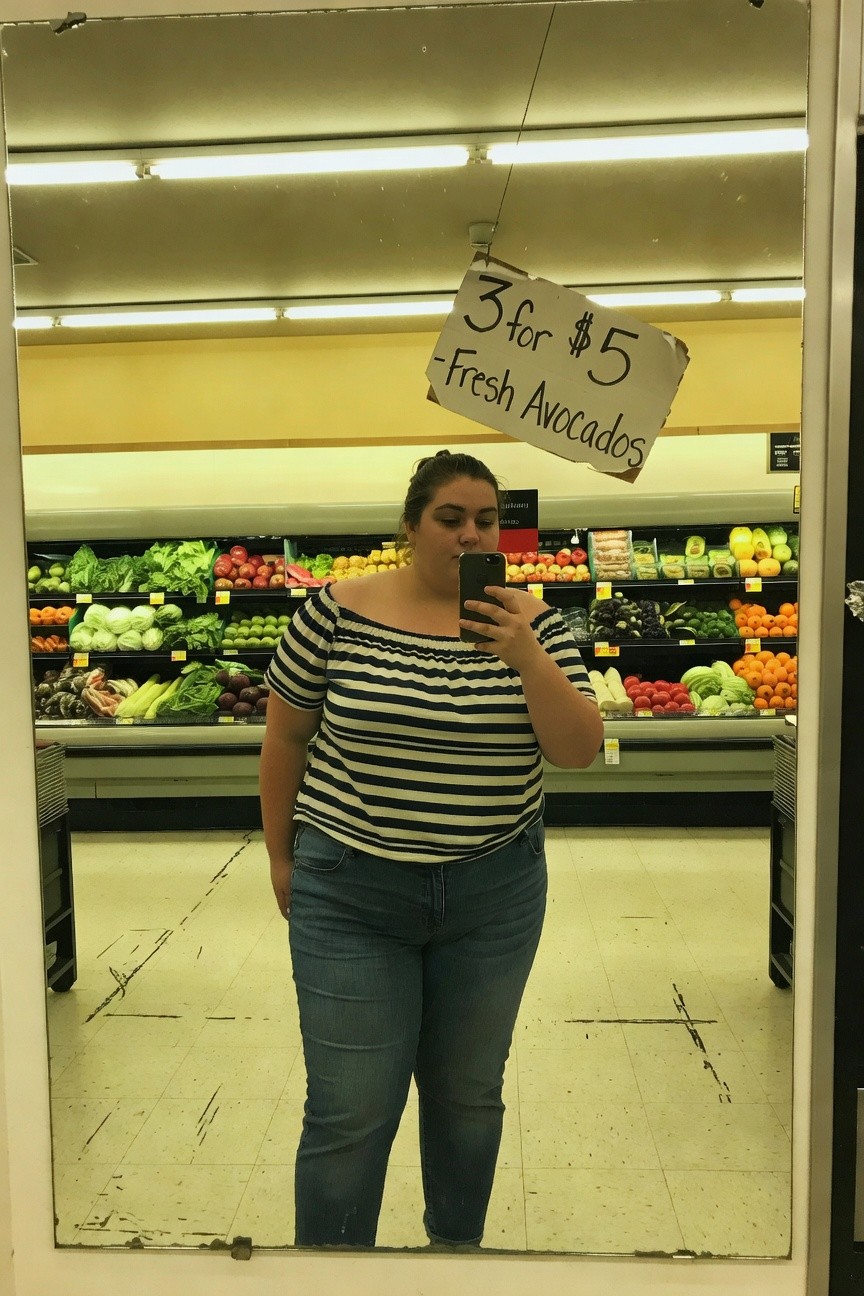 Plus-size woman taking mirror selfie in grocery store wearing black and white striped off-shoulder blouse tucked into blue straight-leg jeans, produce shelves and avocado sale sign in background