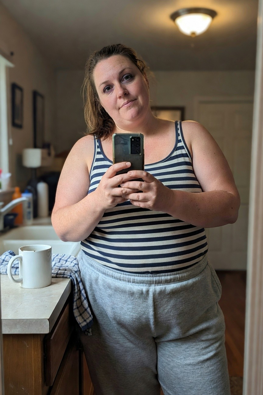 Plus-size woman in a navy and white horizontal striped tank top tucked slightly over gray drawstring jogger sweatpants, standing in a bathroom mirror selfie holding her phone, casual loungewear outfit for summer brunch