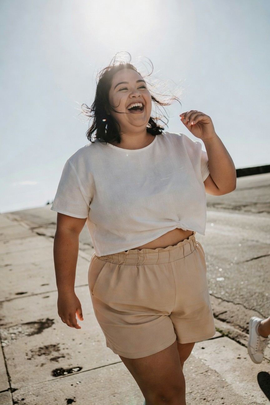 Plus-size woman smiling joyfully while walking in a white short-sleeve cropped twisted top, beige high-waisted pleated shorts, and white sneakers