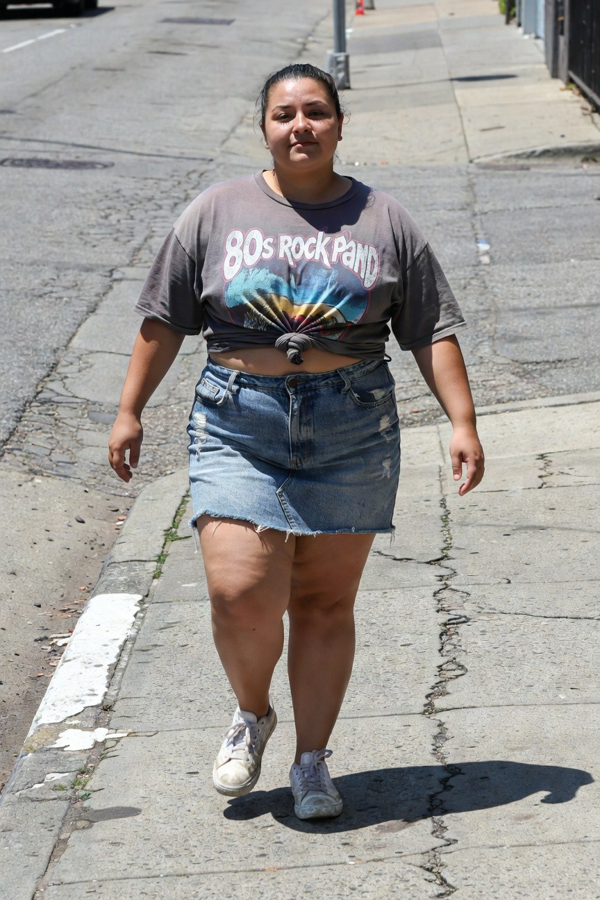 Plus-size woman walking in cropped gray 80s rock band graphic t-shirt tied at the front, high-waisted light wash distressed denim mini skirt, and white sneakers on a city sidewalk