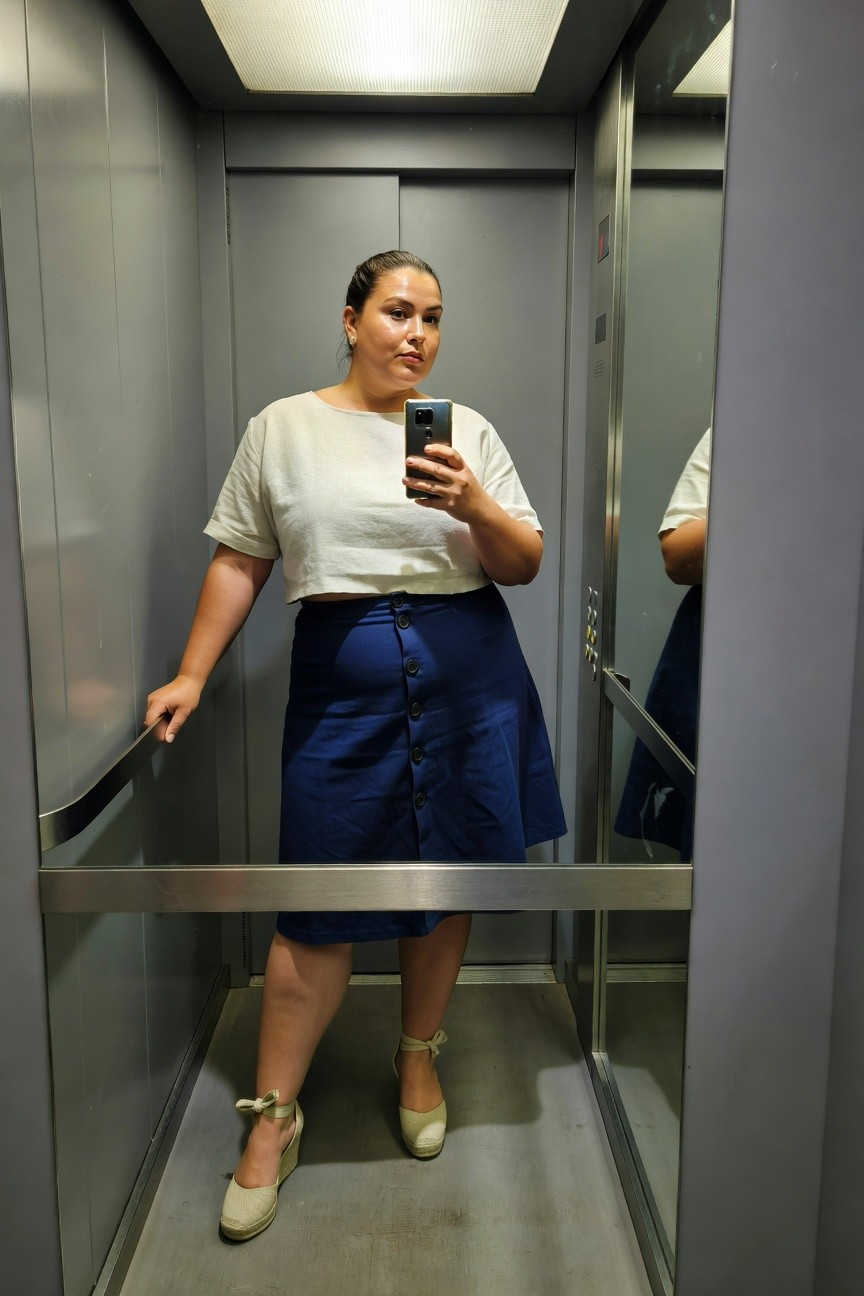 Plus-size woman taking elevator selfie in loose white cropped short-sleeve top, navy blue button-front A-line denim skirt, and beige espadrille wedge sandals with ankle bows, hand on railing.