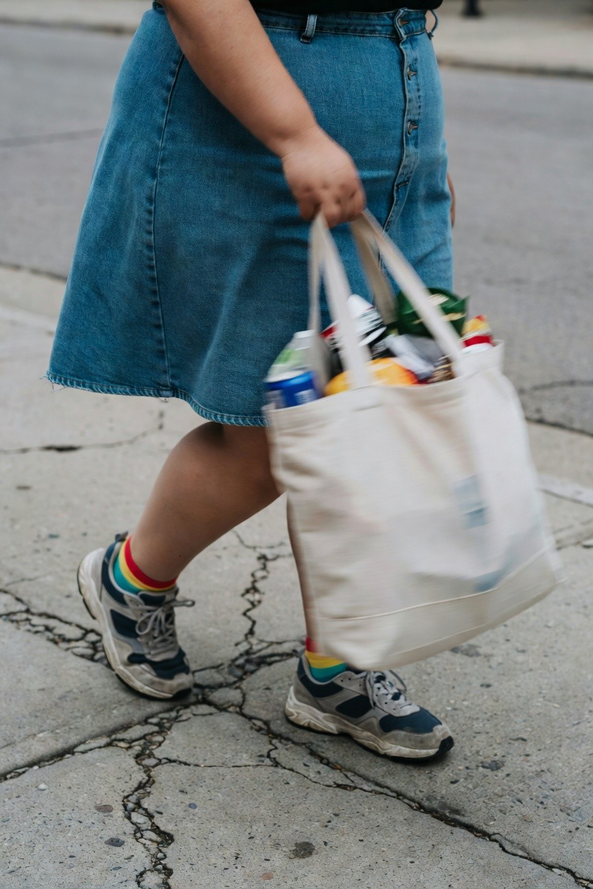 Plus-size woman in high-waisted blue denim skirt, black top, rainbow socks, white and navy chunky sneakers, carrying full white canvas tote bag of groceries while walking on cracked sidewalk