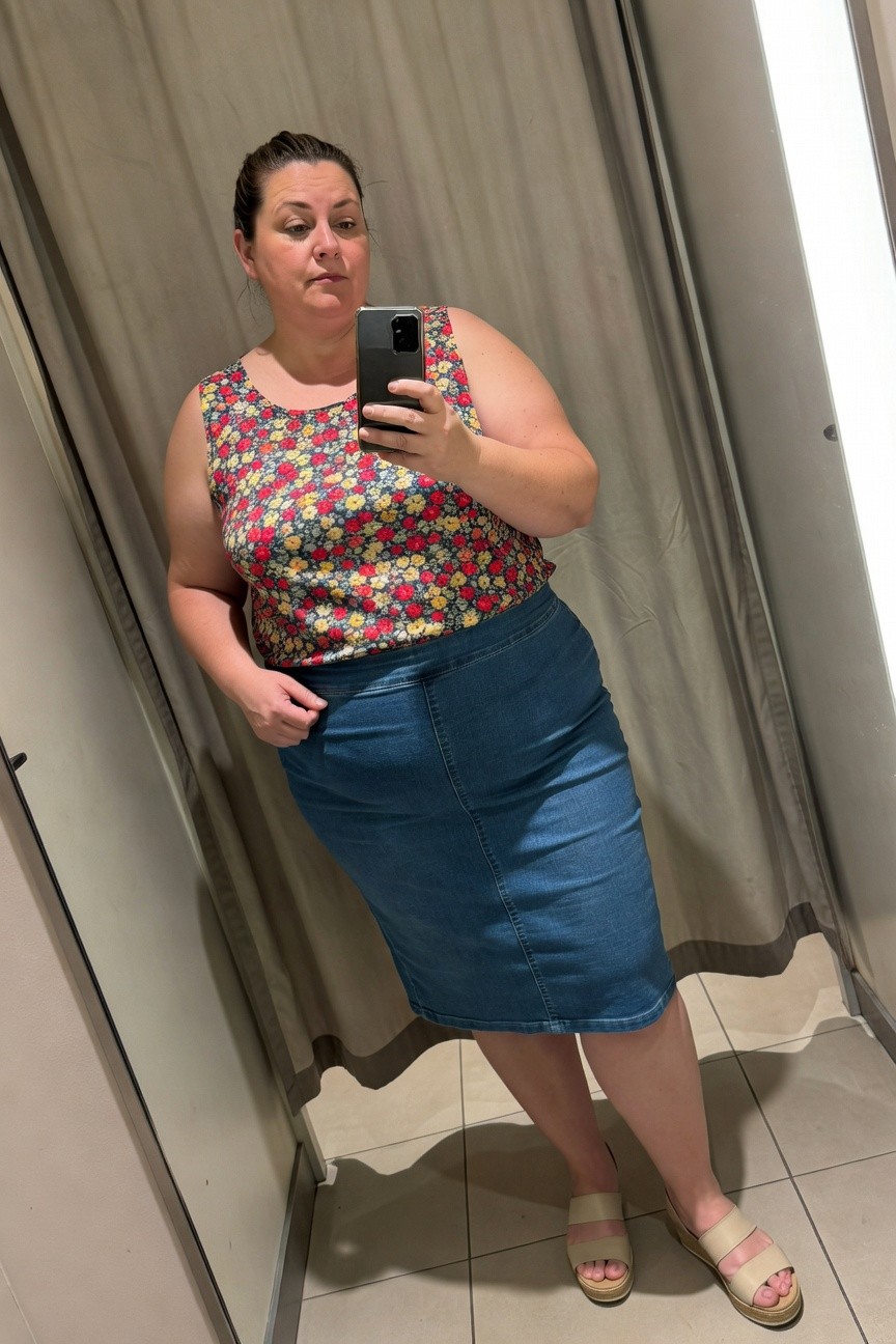 Plus-size woman taking a mirror selfie in a clothing store fitting room, wearing a sleeveless multicolored floral tank top with small flowers in red yellow and white on dark background, high-waisted blue denim midi skirt, and beige strappy flat sandals, one hand in pocket standing sideways