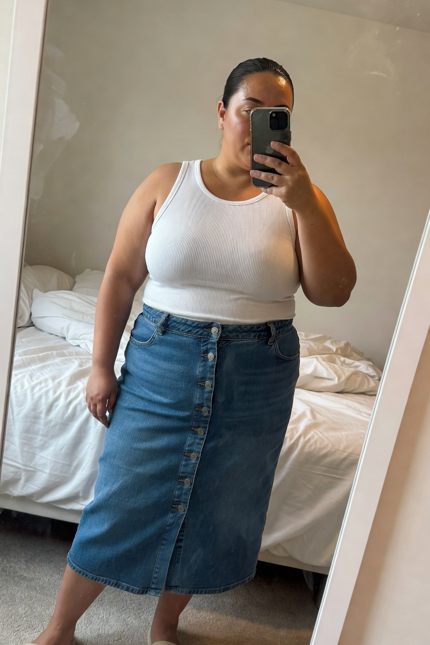 Plus-size woman taking a mirror selfie in a bedroom, wearing a fitted white ribbed sleeveless tank top tucked into a long blue button-front denim skirt with straight silhouette, paired with beige flat espadrilles, natural indoor lighting