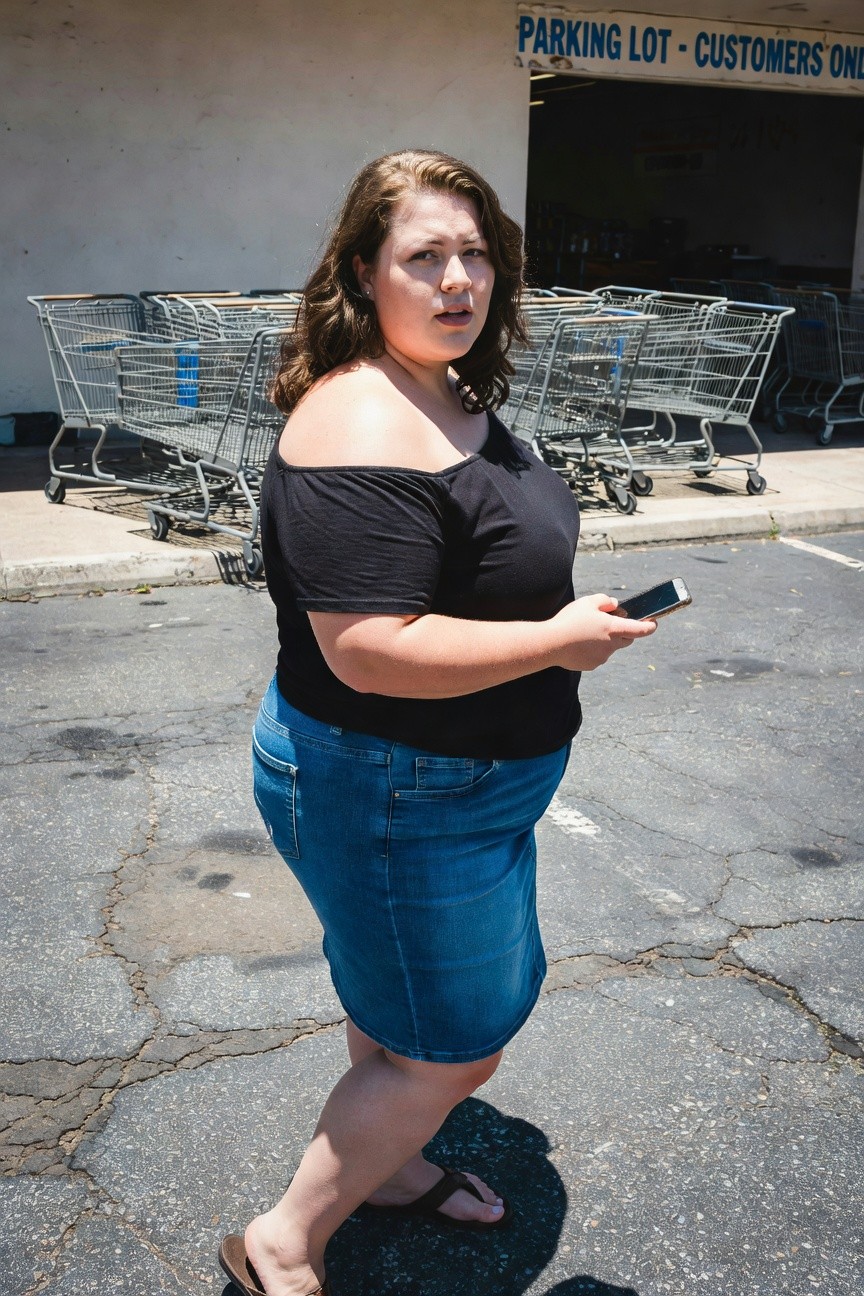 Plus-size woman standing in parking lot wearing black off-shoulder top, high-waisted blue denim skirt, brown flip-flops, holding smartphone, surrounded by shopping carts