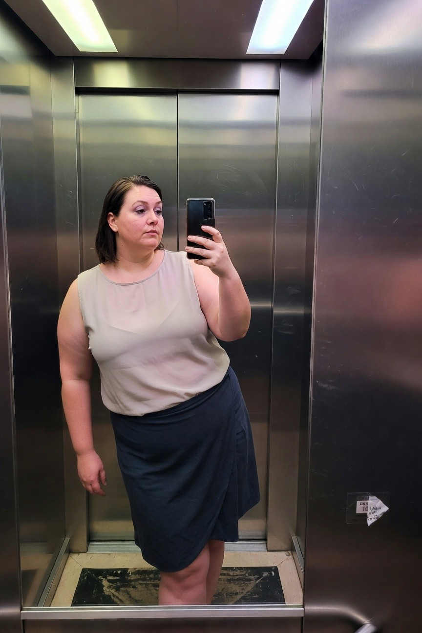 Plus-size woman in elevator selfie wearing light beige sleeveless chiffon top and dark navy knee-length wrap skirt, one hand on hip, phone in other
