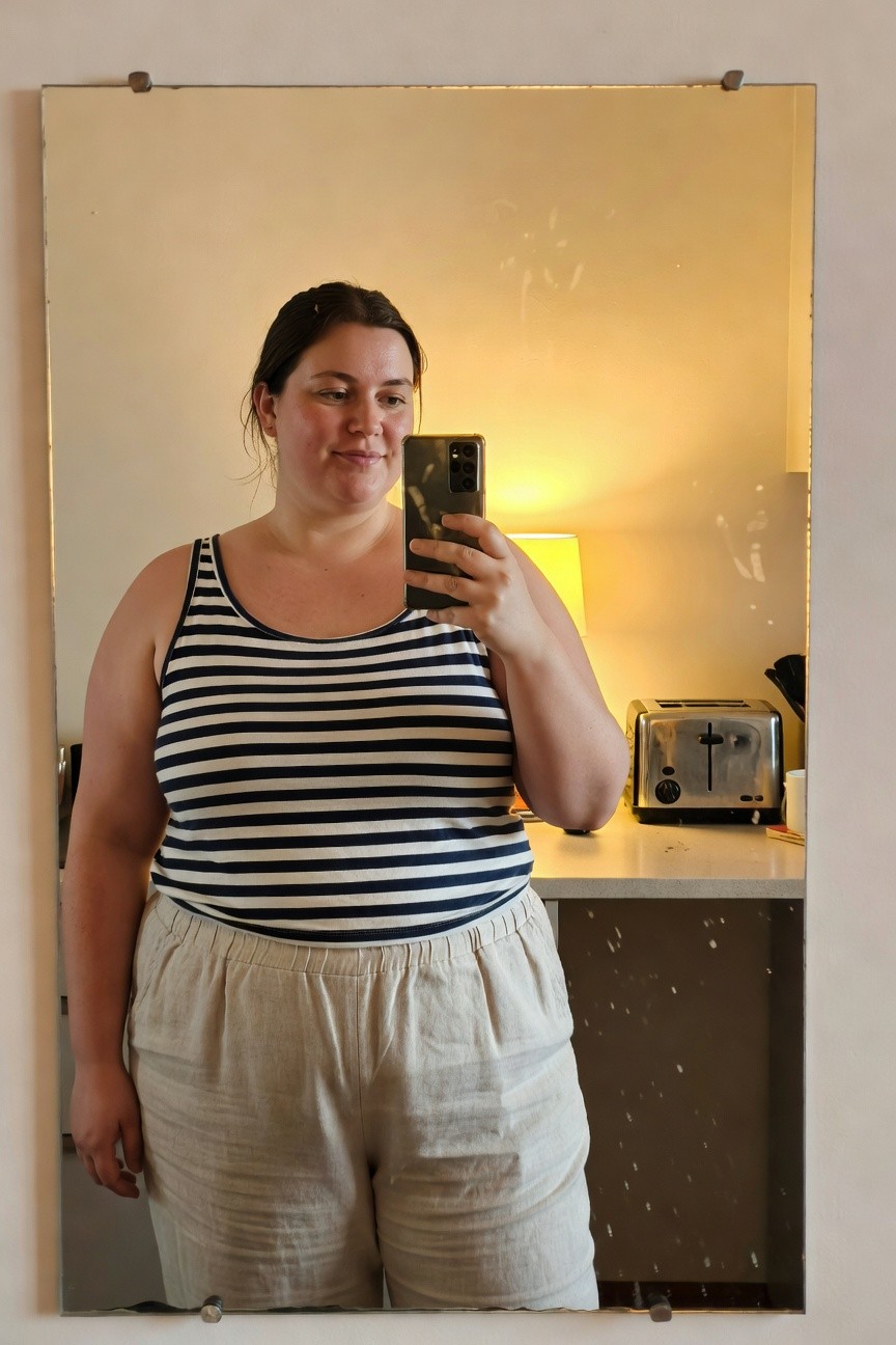 Mirror selfie of plus-size woman in navy and white horizontal striped sleeveless tank top tucked slightly into high-waisted beige linen wide-leg pants, standing in kitchen area with toaster and lamp in background