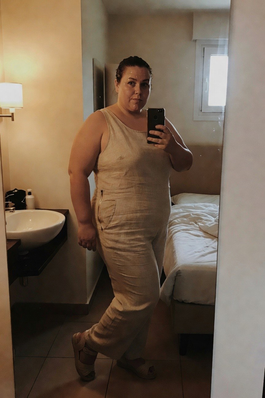 Plus-size woman in a tan sleeveless linen jumpsuit with wide legs and front zipper, posing in front of a bathroom mirror while holding her phone for a selfie, wearing beige wedge sandals, hotel room setting with sink and bed visible.