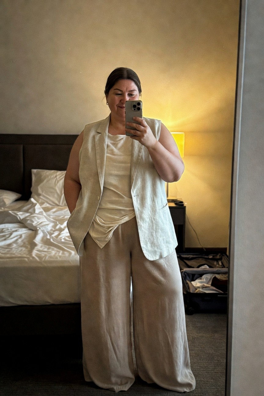 Plus-size woman in cream linen sleeveless blazer vest over white tank top and matching wide-leg linen pants, hands in pockets, taking mirror selfie in beige hotel room with bed and lamp in background