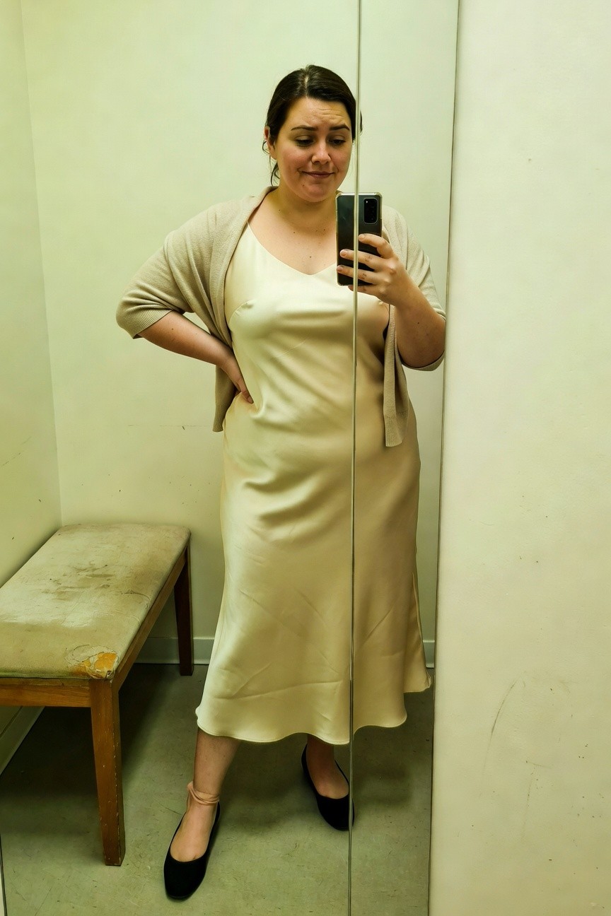 Plus-size woman in a cream satin midi slip dress with matching open beige cardigan, hands on hips, wearing black ballet flats with ankle straps, selfie in dressing room mirror.