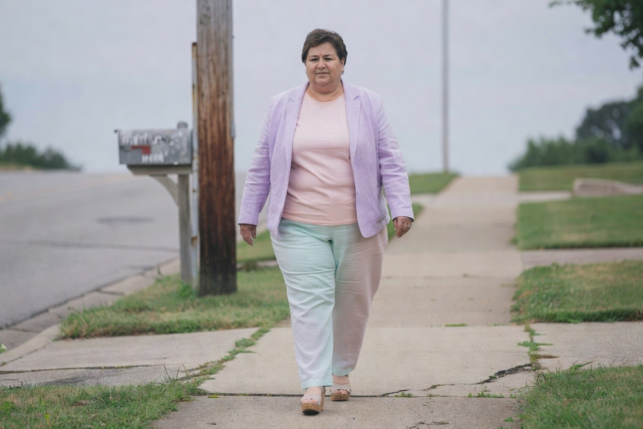 Plus-size woman walking on sidewalk in light purple linen blazer over pink top, colorblock wide-leg pants with one mint green leg and one pale leg, tan wedge sandals, silver ring on finger