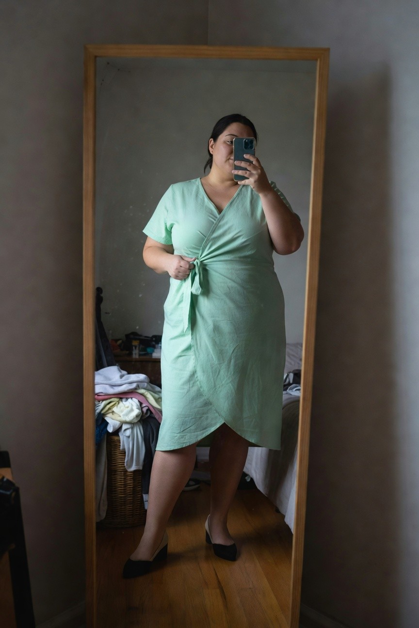 Plus-size woman in a light green short-sleeve wrap dress with waist tie, posing in front of a mirror, wearing black low-heel pumps, bedroom visible in reflection