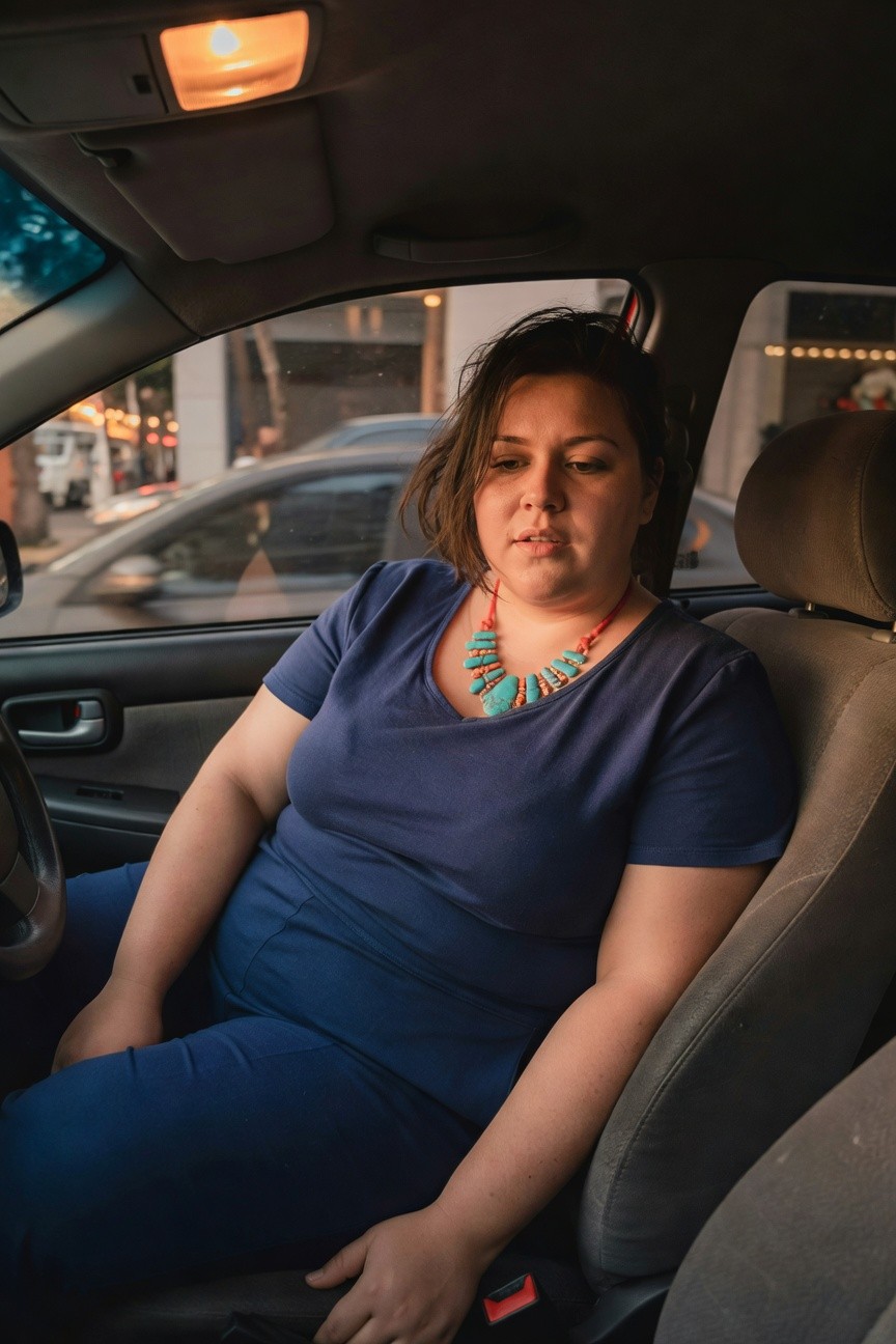 Plus-size woman wearing a navy blue short-sleeved V-neck top tucked into matching straight-leg pants, accented by a chunky turquoise and coral beaded necklace, seated in a car driver's seat