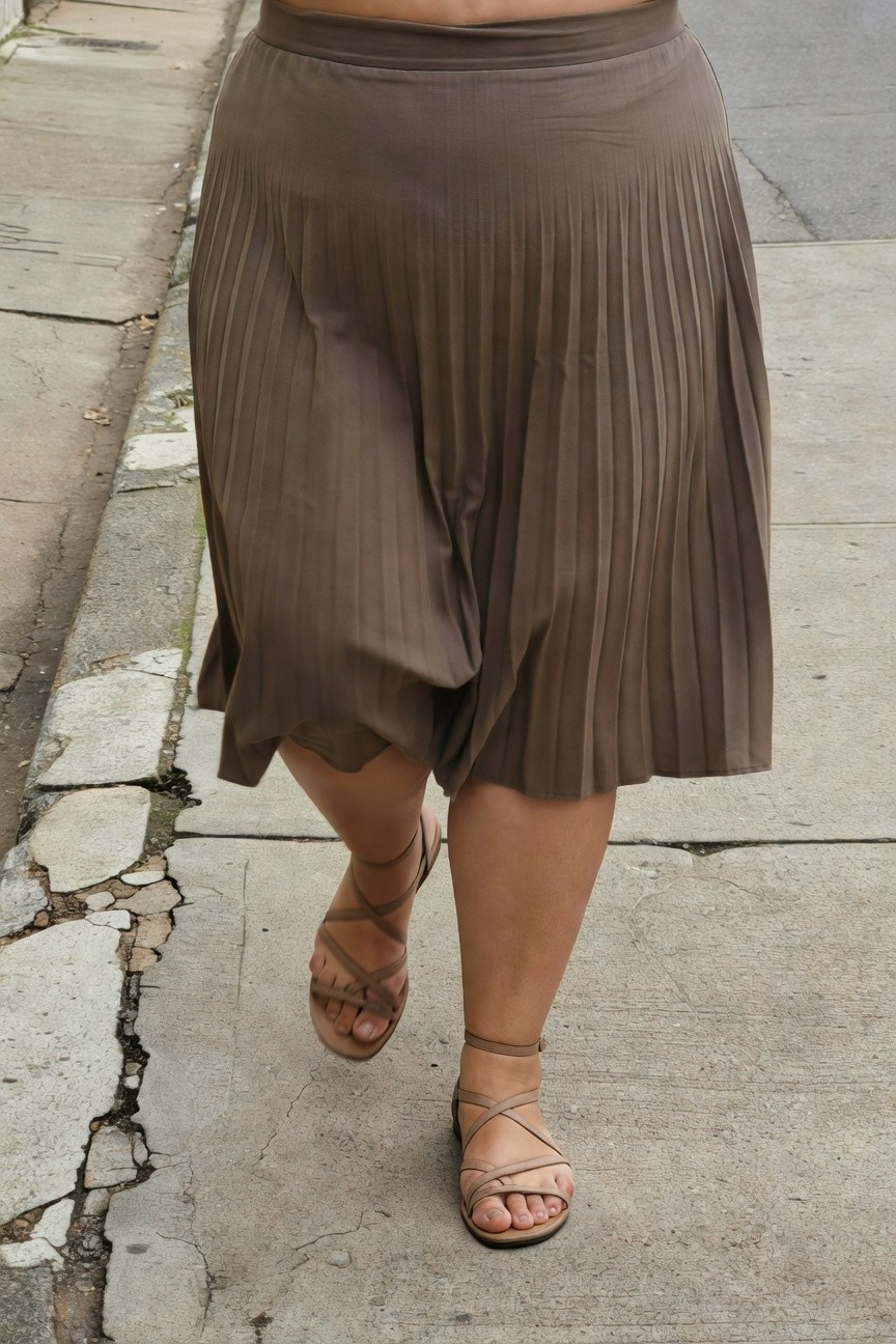 Plus-size woman walking on sidewalk in high-waisted taupe pleated midi skirt and beige strappy flat sandals, emphasizing flowy skirt movement and leg exposure