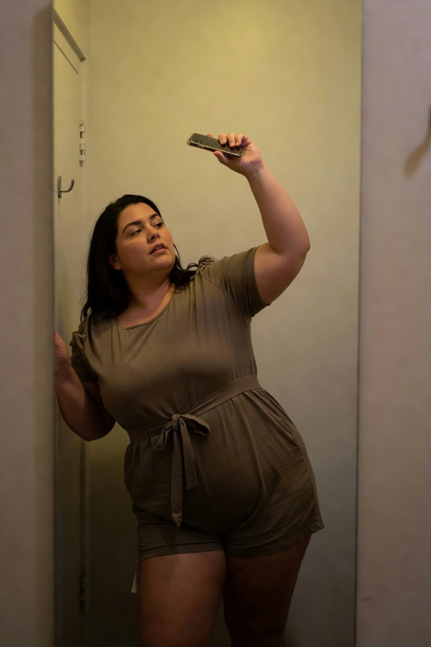 Plus-size woman with long dark hair taking a selfie in a hallway, wearing a short-sleeved taupe belted romper that accentuates her curves