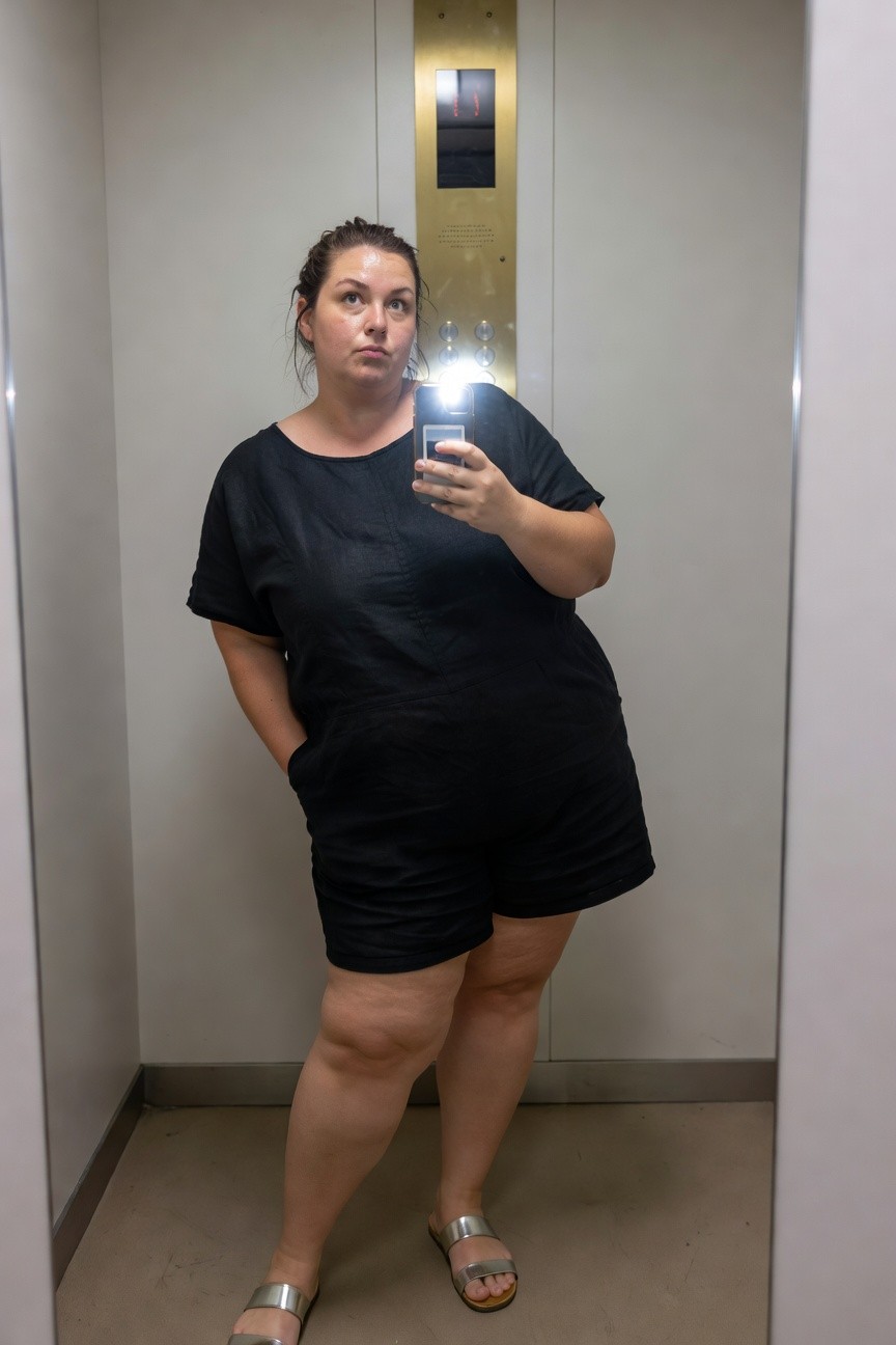Plus-size woman in black linen romper with short sleeves, shorts, and side pockets, posing casually with hand in pocket while holding phone for mirror selfie, wearing silver strappy sandals in elevator