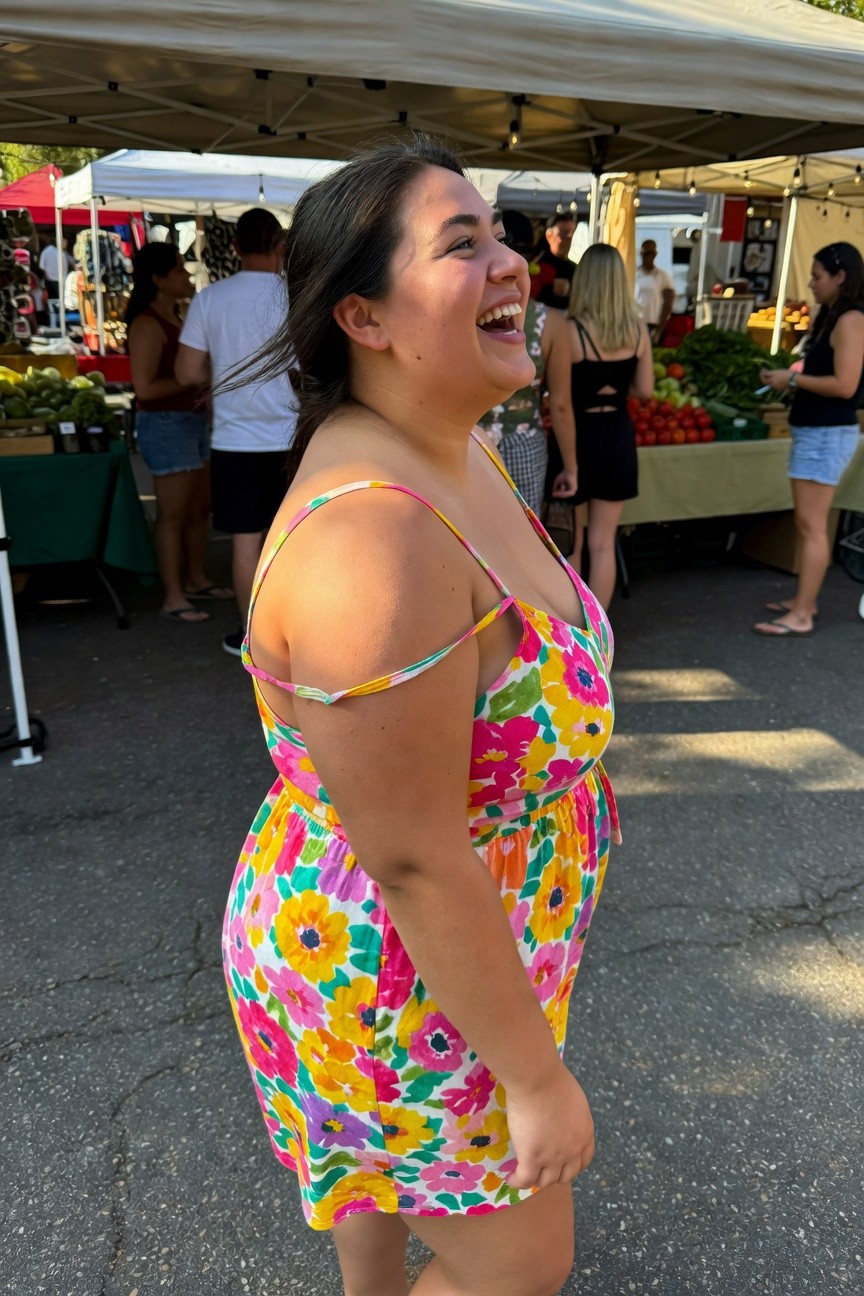 Plus-size woman in profile smiling while wearing a vibrant multicolored floral print spaghetti-strap romper with a low neckline, cinched waist, and short hemline at a farmers market
