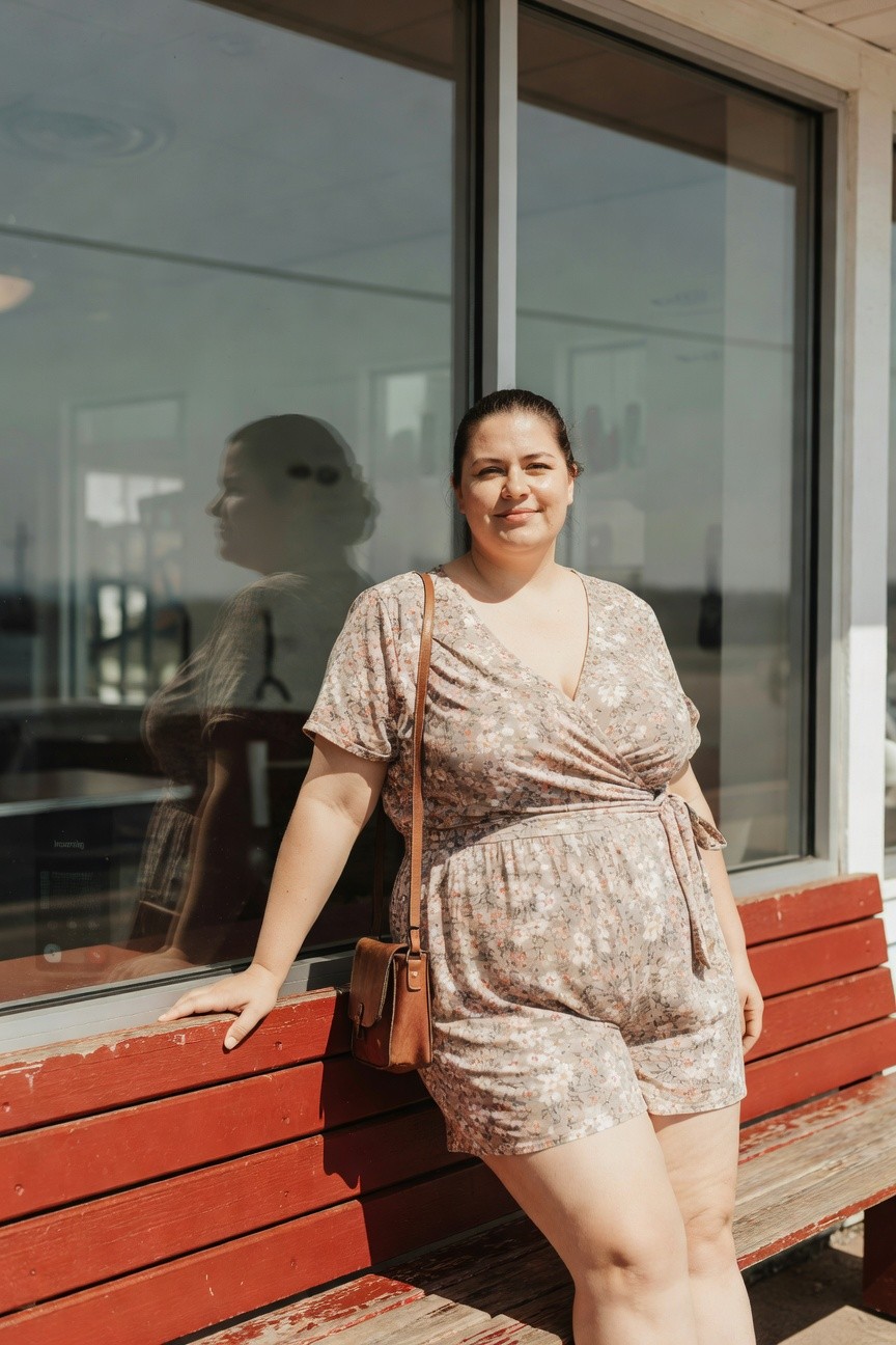 Plus-size woman in a beige floral wrap romper with short sleeves and shorts, tan leather crossbody bag, leaning against a window with hand on red bench